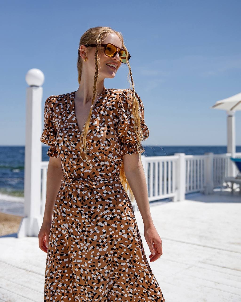 Woman in a patterned dress with braided hair standing on a beach deck.