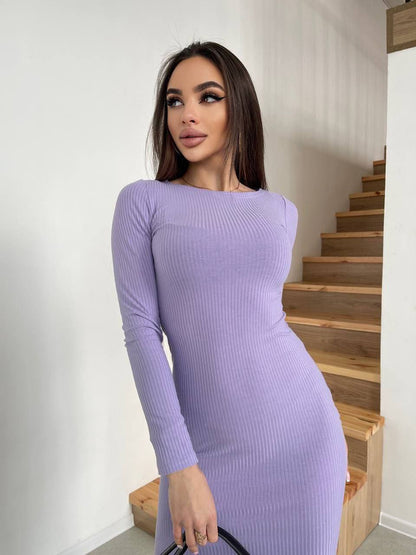 Woman wearing a purple ribbed dress standing in a home setting with wooden stairs.