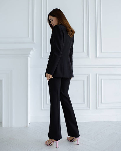Woman wearing a black suit standing against a white paneled wall.