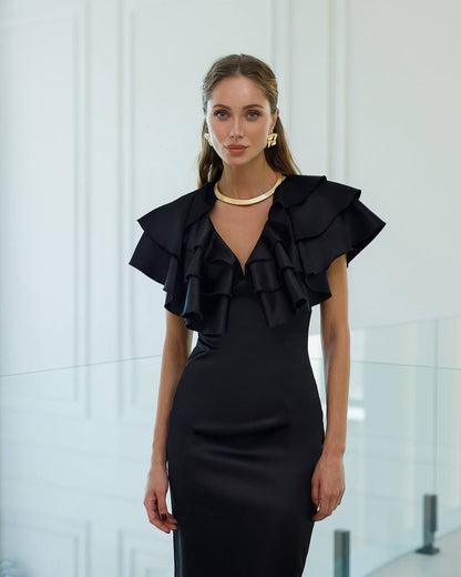 Woman wearing a black dress with ruffled details against a white wall.