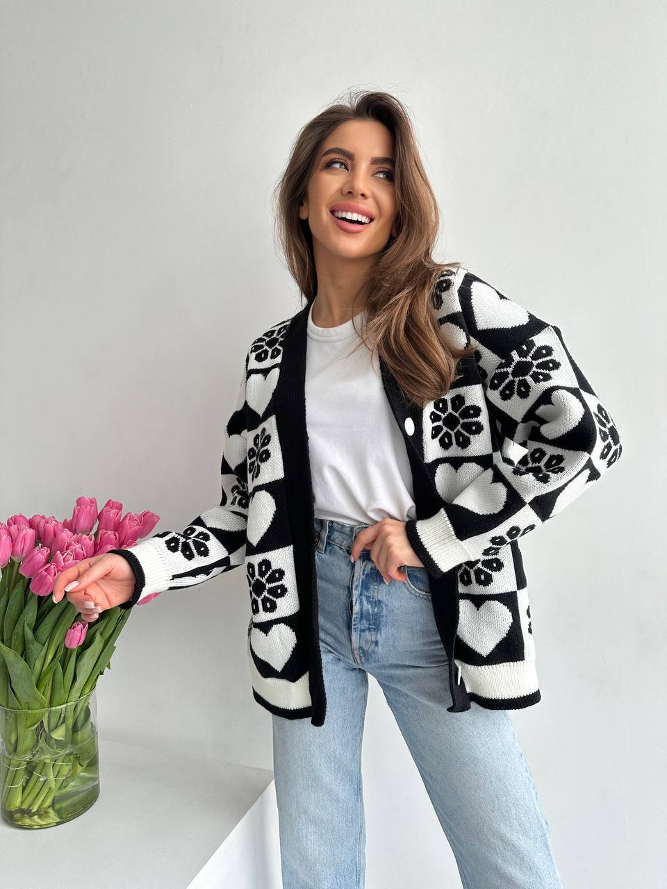 Woman wearing a black and white patterned cardigan holding pink tulips against a plain background