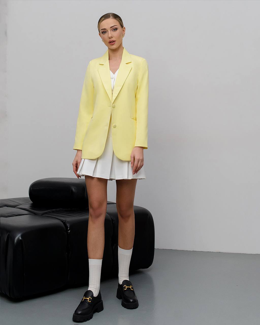 A model stands in a studio setting wearing a pale yellow blazer over a white top and white pleated skirt, with white socks and black loafers. A black leather bench is visible against a light gray wall and floor.