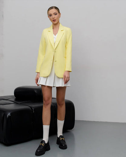 A model stands in a studio setting wearing a pale yellow blazer over a white top and white pleated skirt, with white socks and black loafers. A black leather bench is visible against a light gray wall and floor.