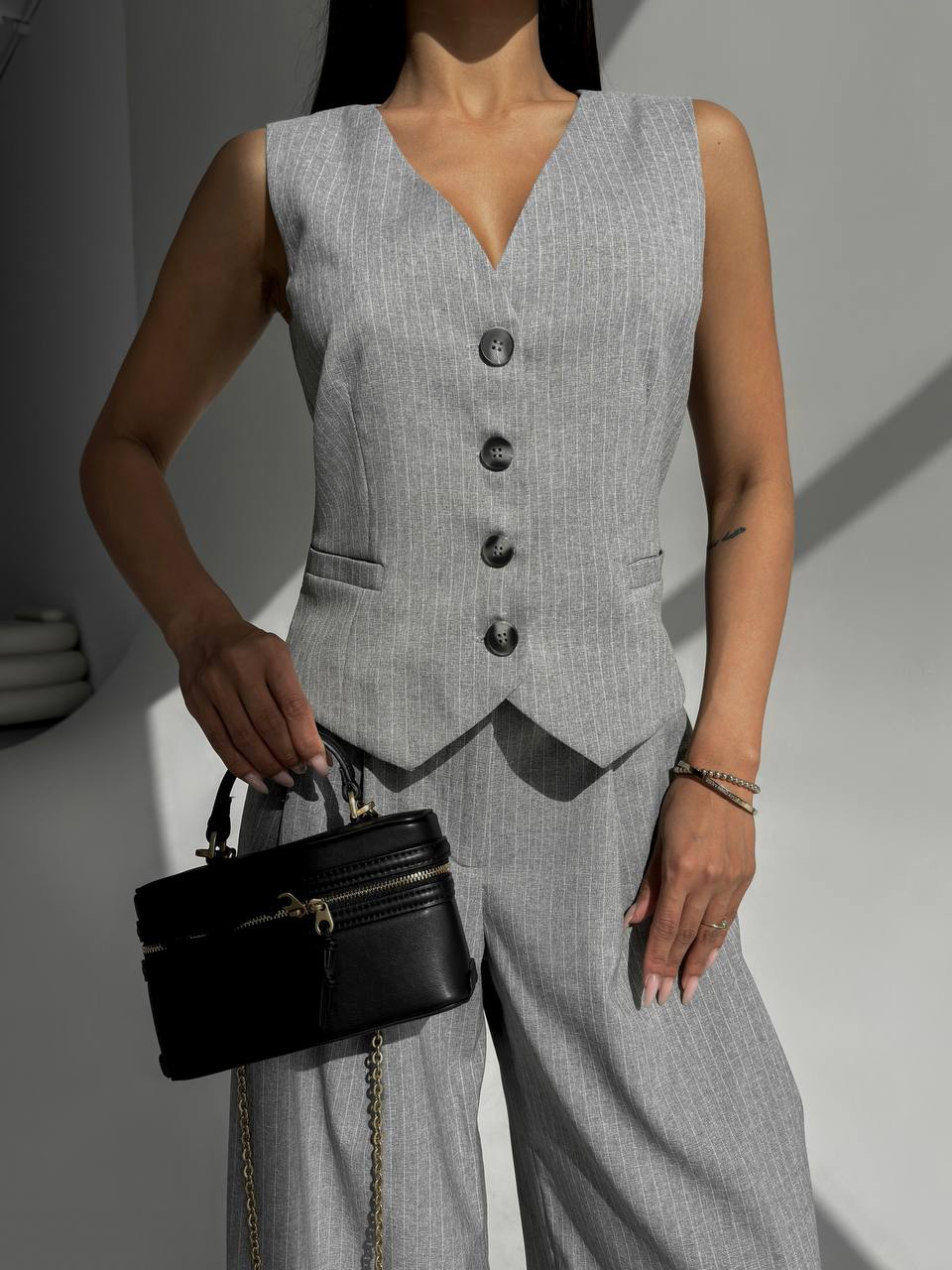 Person wearing a gray vest and holding a black handbag against a neutral background