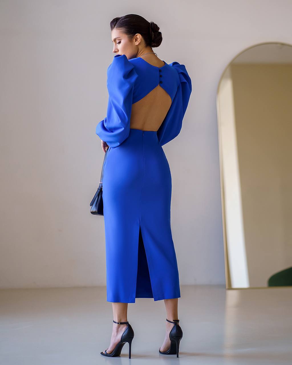 Woman wearing a blue dress with an open back, standing in a room with a mirror.