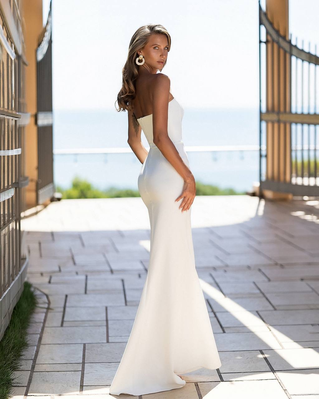 A woman wearing a strapless white mermaid-style evening gown stands on a tiled walkway outdoors, with a gate, ocean, and bright sky in the background.