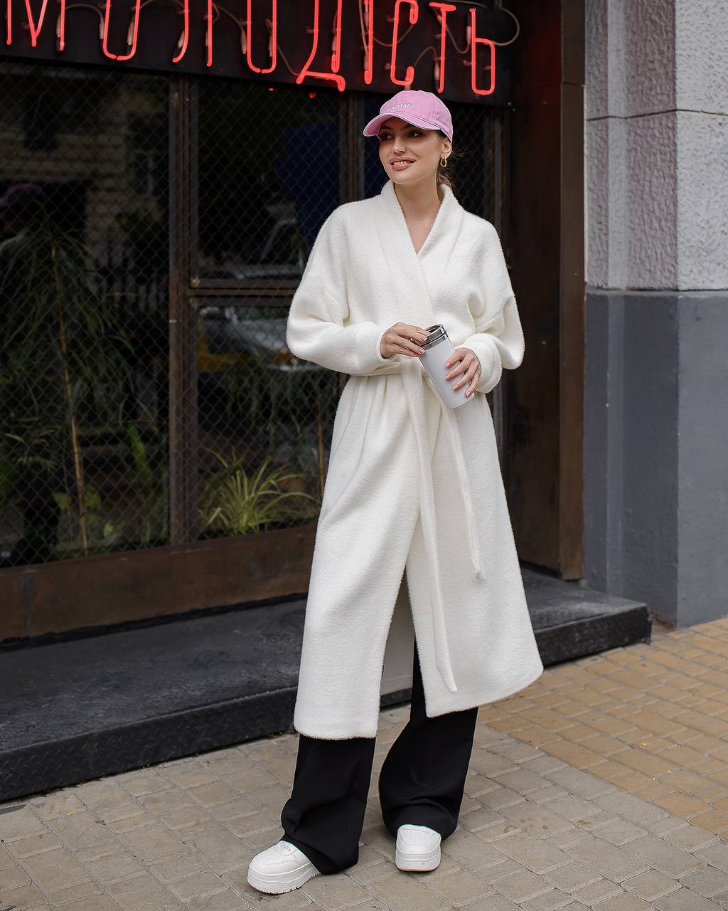 Person wearing a white coat and black pants standing in front of a building with neon signage.