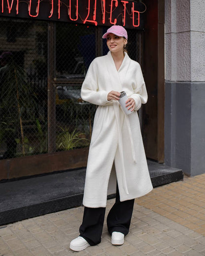Person wearing a white coat and black pants standing in front of a building with neon signage.