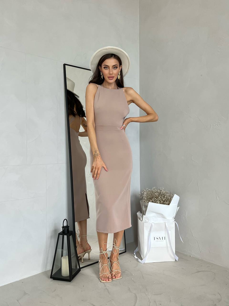 Woman in a beige dress standing in front of a mirror, wearing a white hat.