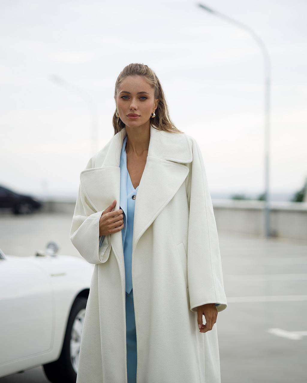 Woman wearing a long white coat over a blue outfit in an outdoor setting.