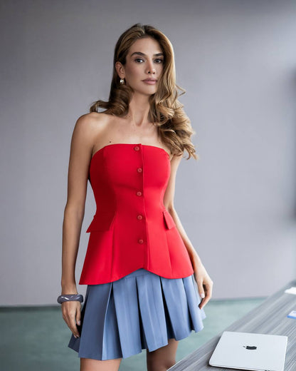 A woman modeling a bright red strapless corset-style top with front buttons, paired with a light blue pleated skirt, standing in a studio setting.