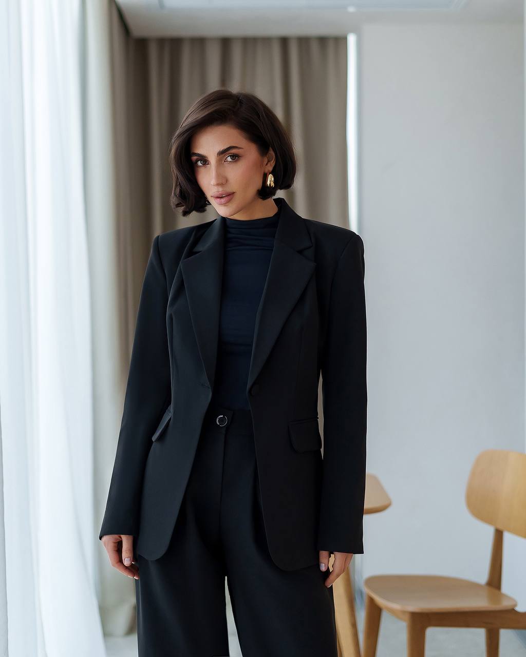 Woman wearing a black suit standing in a room with curtains and a chair.