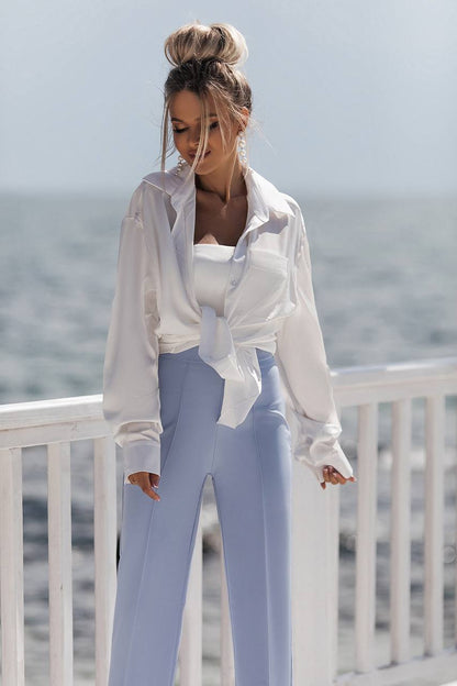 Woman wearing a white blouse and light blue pants standing on a balcony with a blurred ocean background