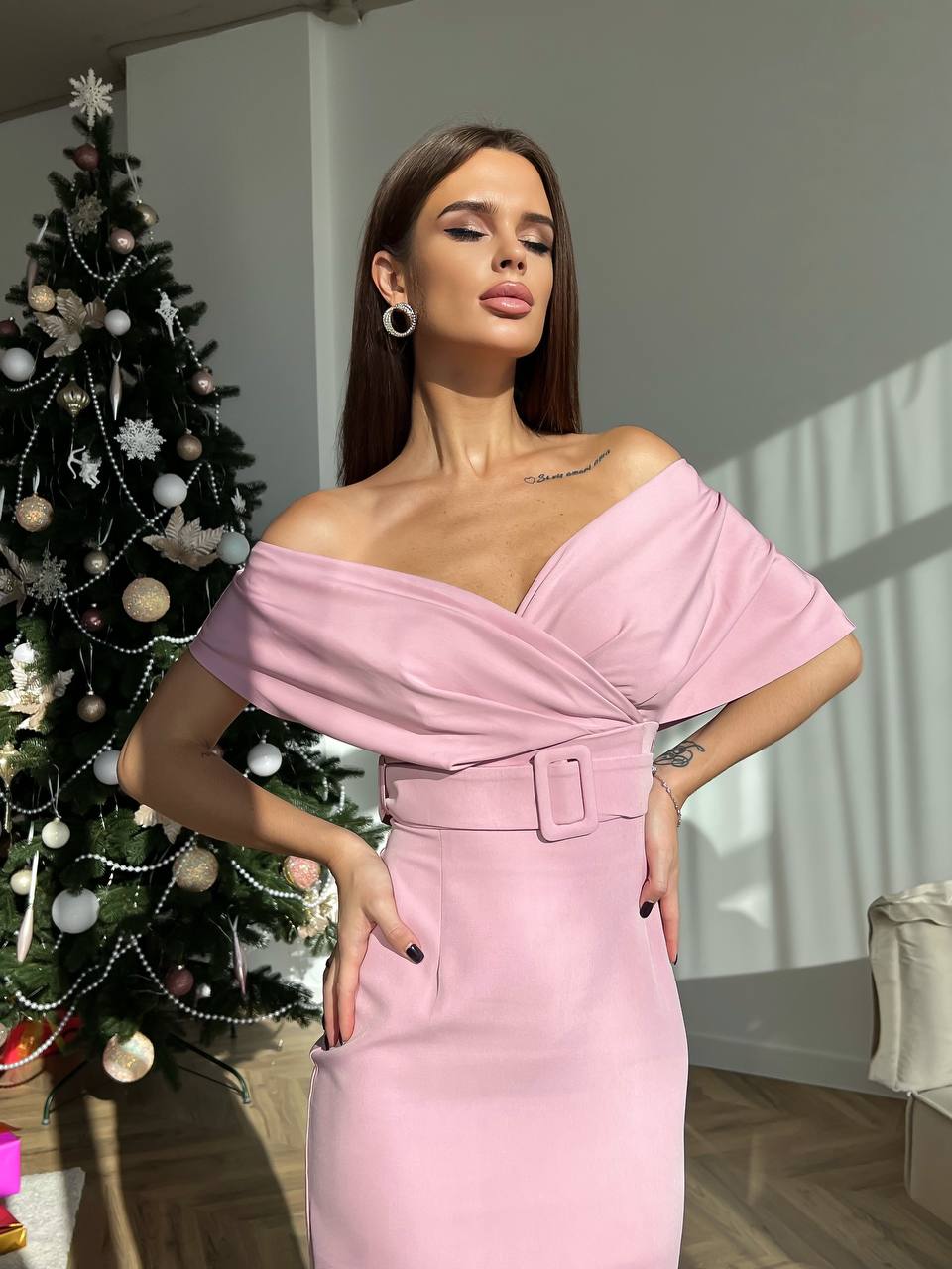 Woman in a pink off-shoulder dress standing next to a decorated Christmas tree indoors.