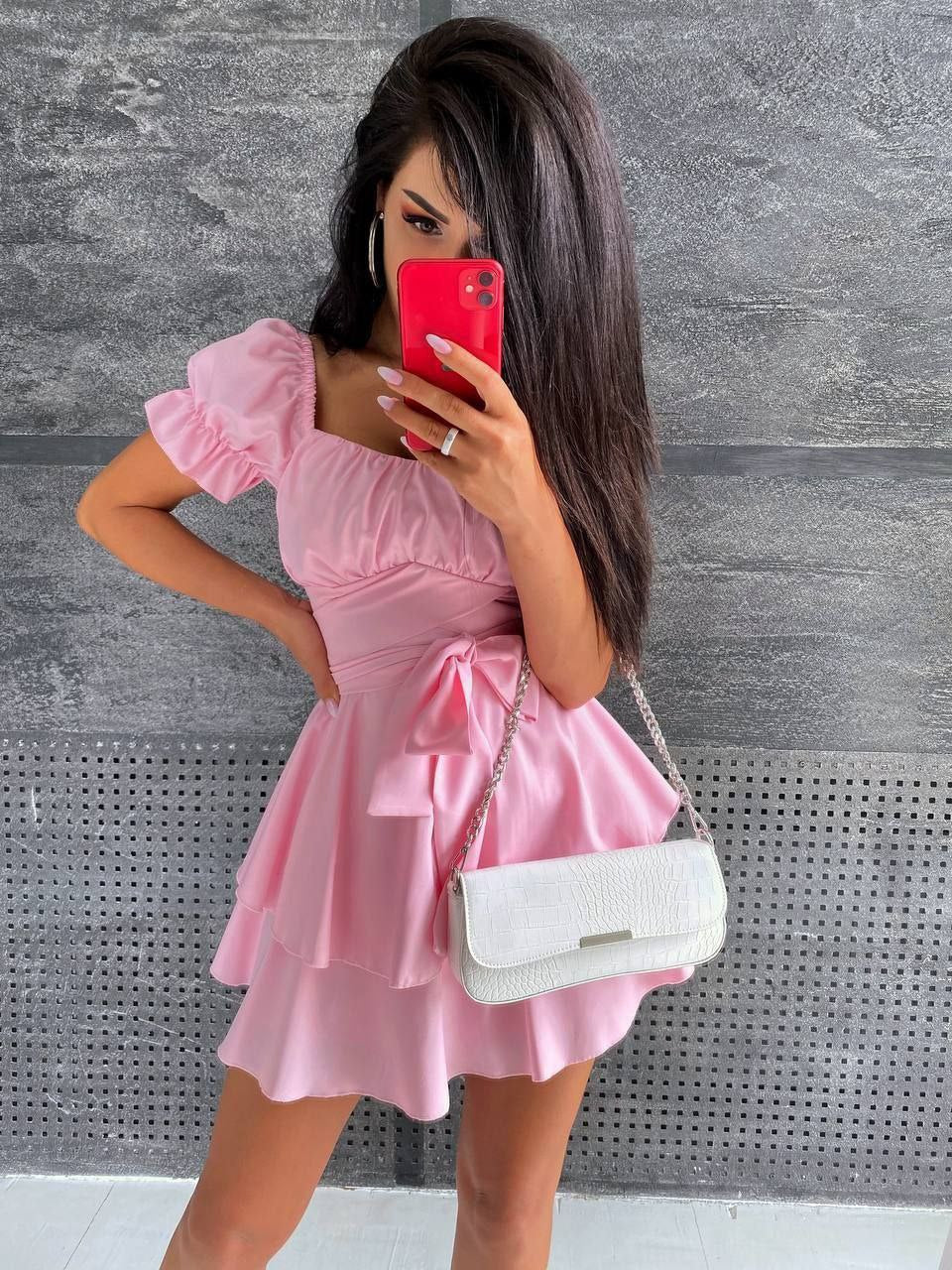 Woman in a pink dress holding a white handbag against a gray wall.
