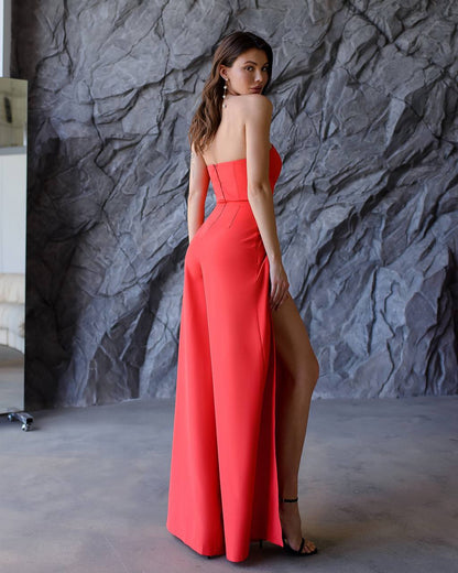 Woman in a strapless red gown with a high slit standing against a textured gray wall.
