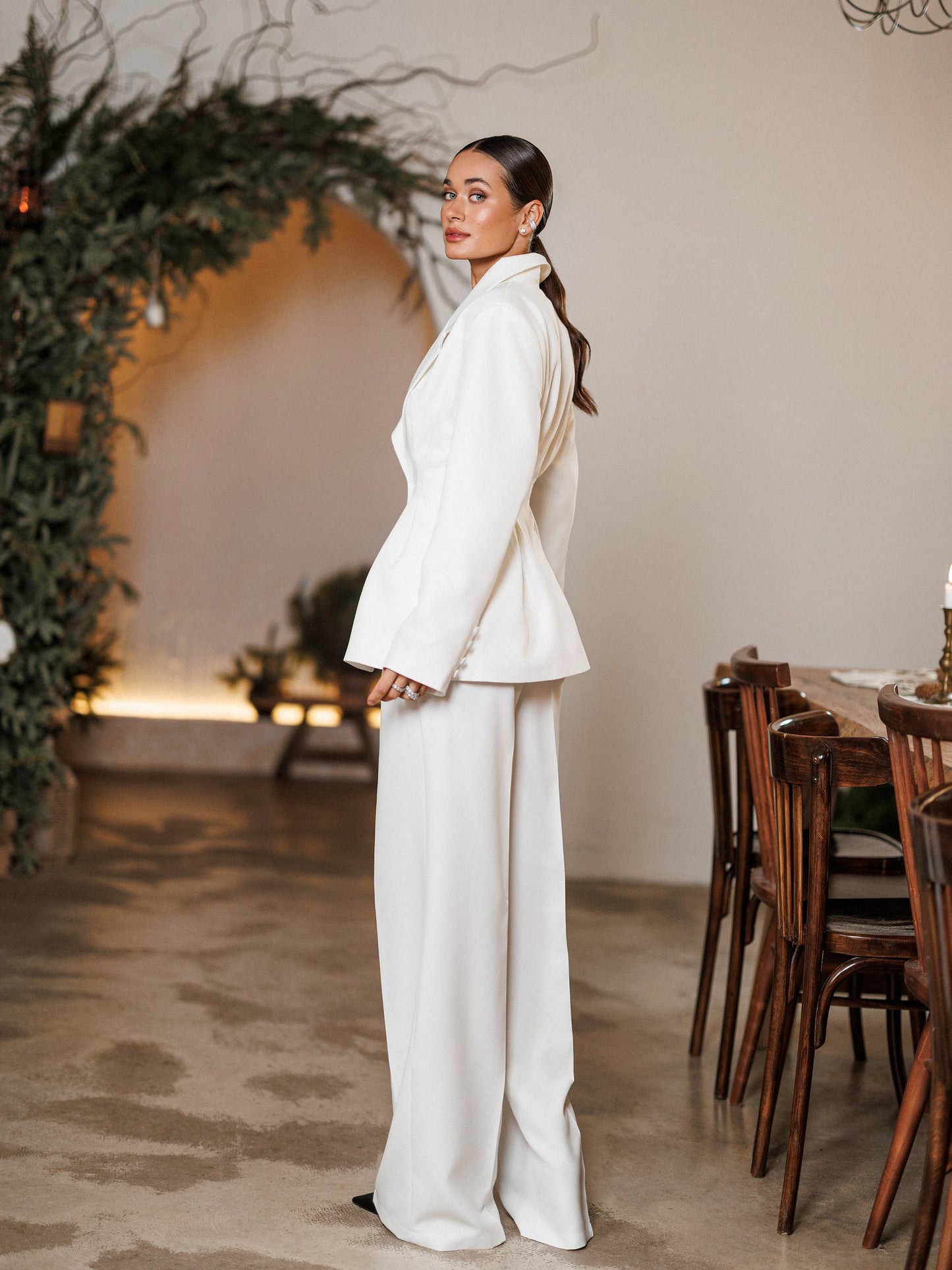 Woman in a white suit standing in a room with wooden chairs and decorative plants.