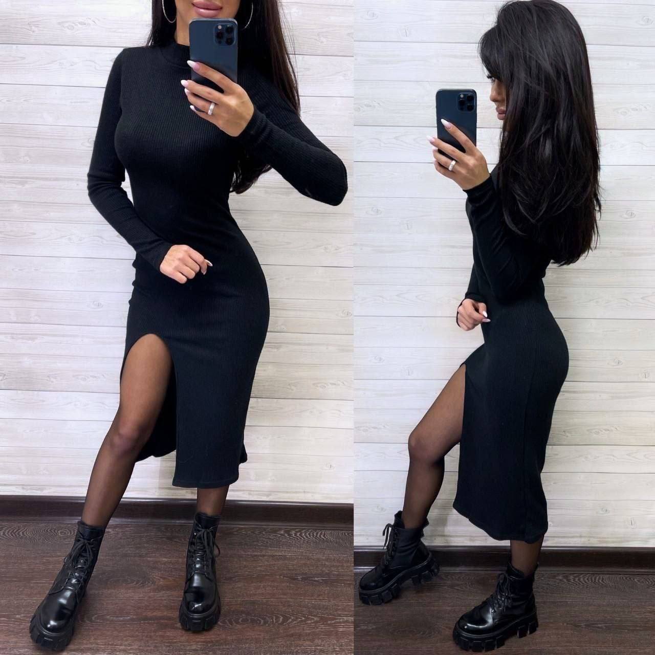 Woman wearing a black dress with a slit, standing against a wooden panel background.