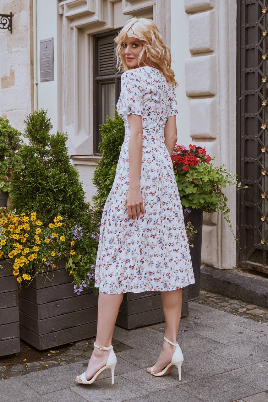 Woman in a floral dress standing on a street with decorative plants and flowers.