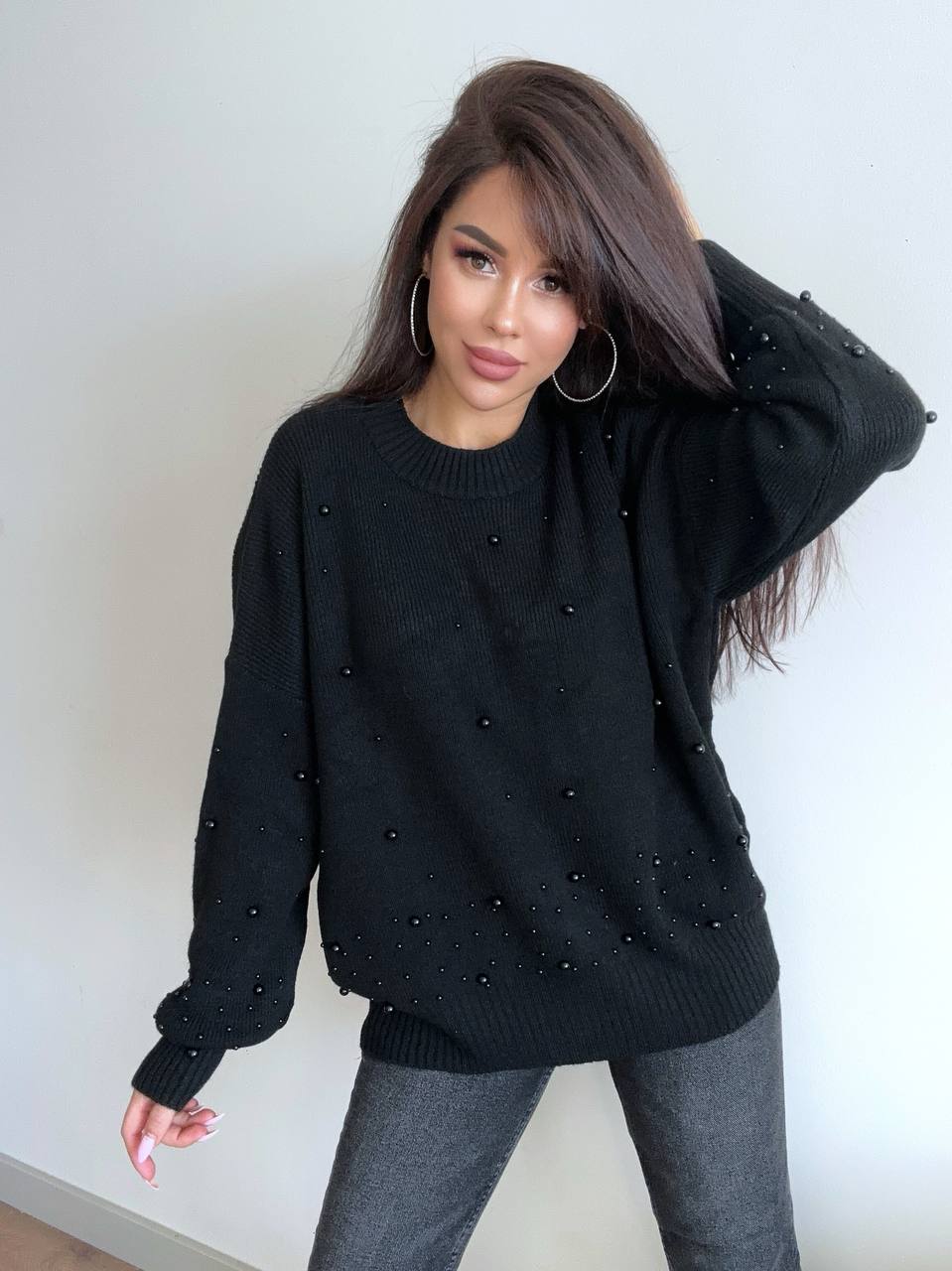 Woman wearing a black sweater with pearl embellishments against a white wall.