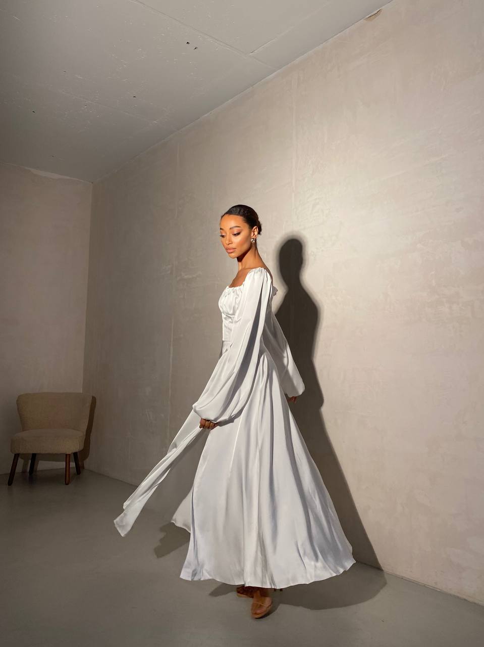 Woman in a white dress standing in a minimalistic room with a chair in the corner.