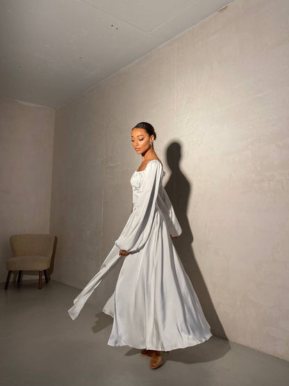 Woman in a white dress standing in a minimalistic room with a chair in the corner.