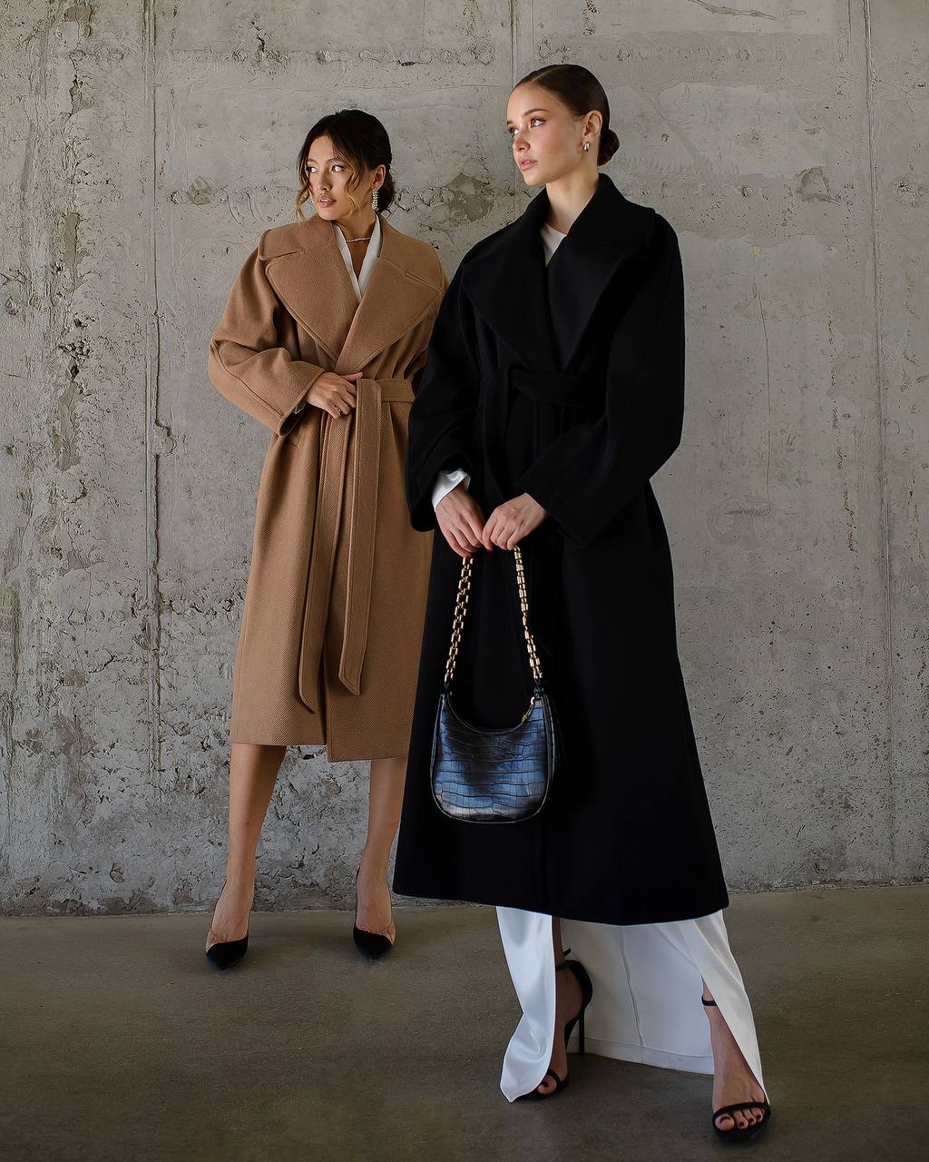 Two women wearing long coats standing against a plain wall.