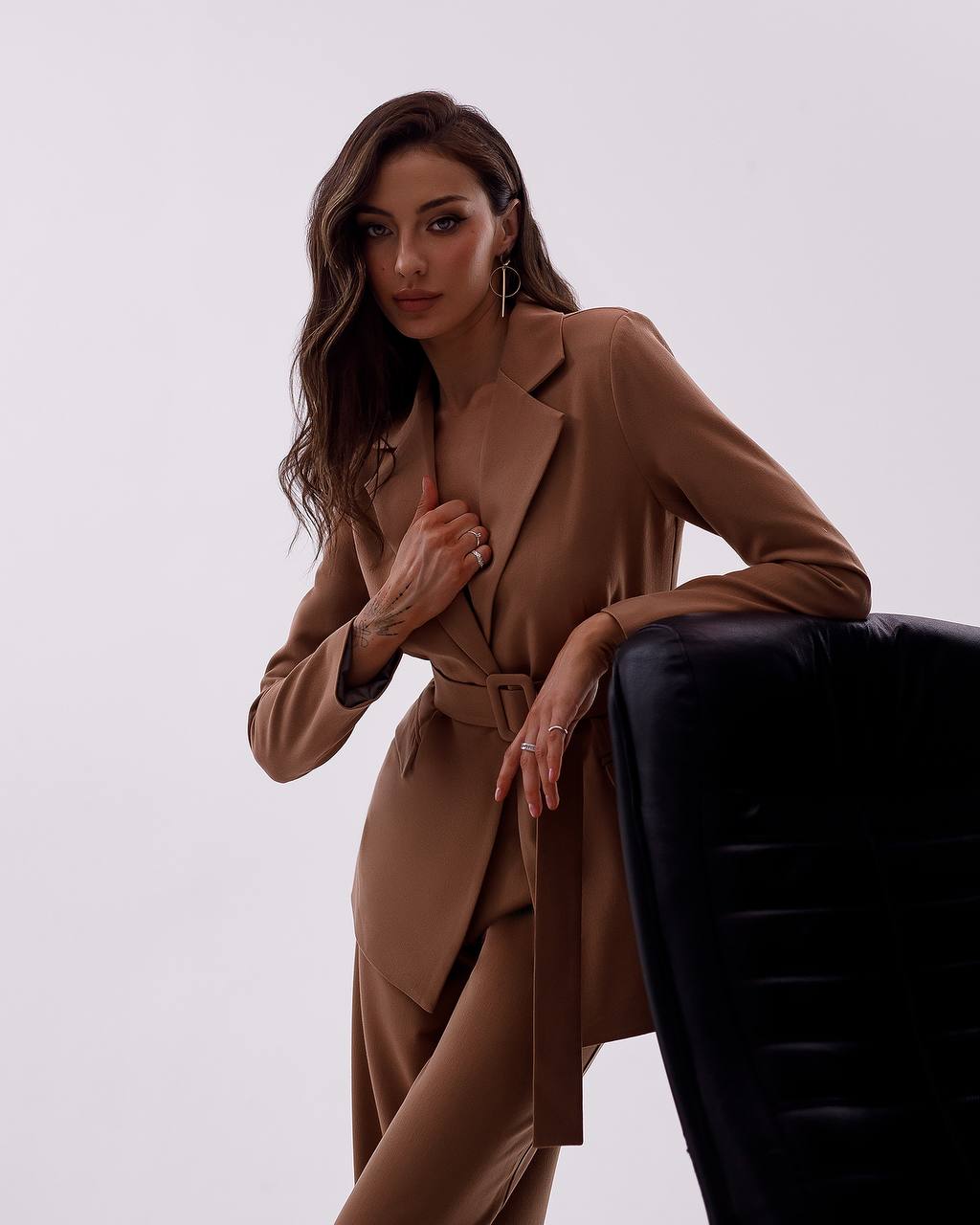 Woman wearing a brown coat leaning against a black chair on a white background
