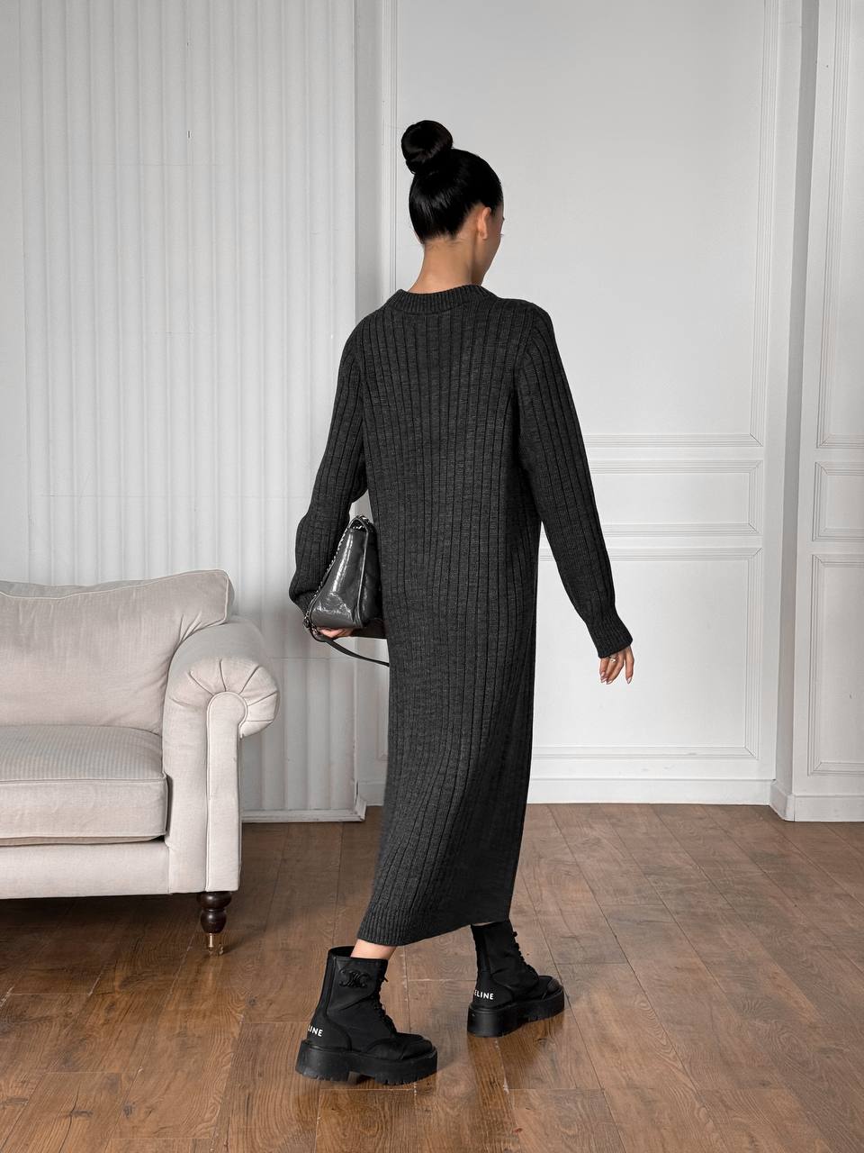 Woman in a long dark gray ribbed dress standing in a room with a sofa and white walls.