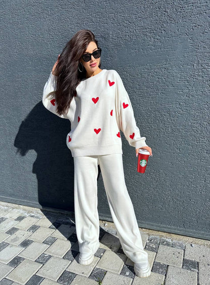 Person wearing a white sweater with red heart patterns and white pants, holding a red cup against a gray wall.