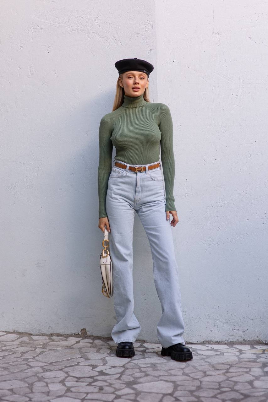 Person wearing a green turtleneck, light blue jeans, black beret, and black shoes against a white wall.
