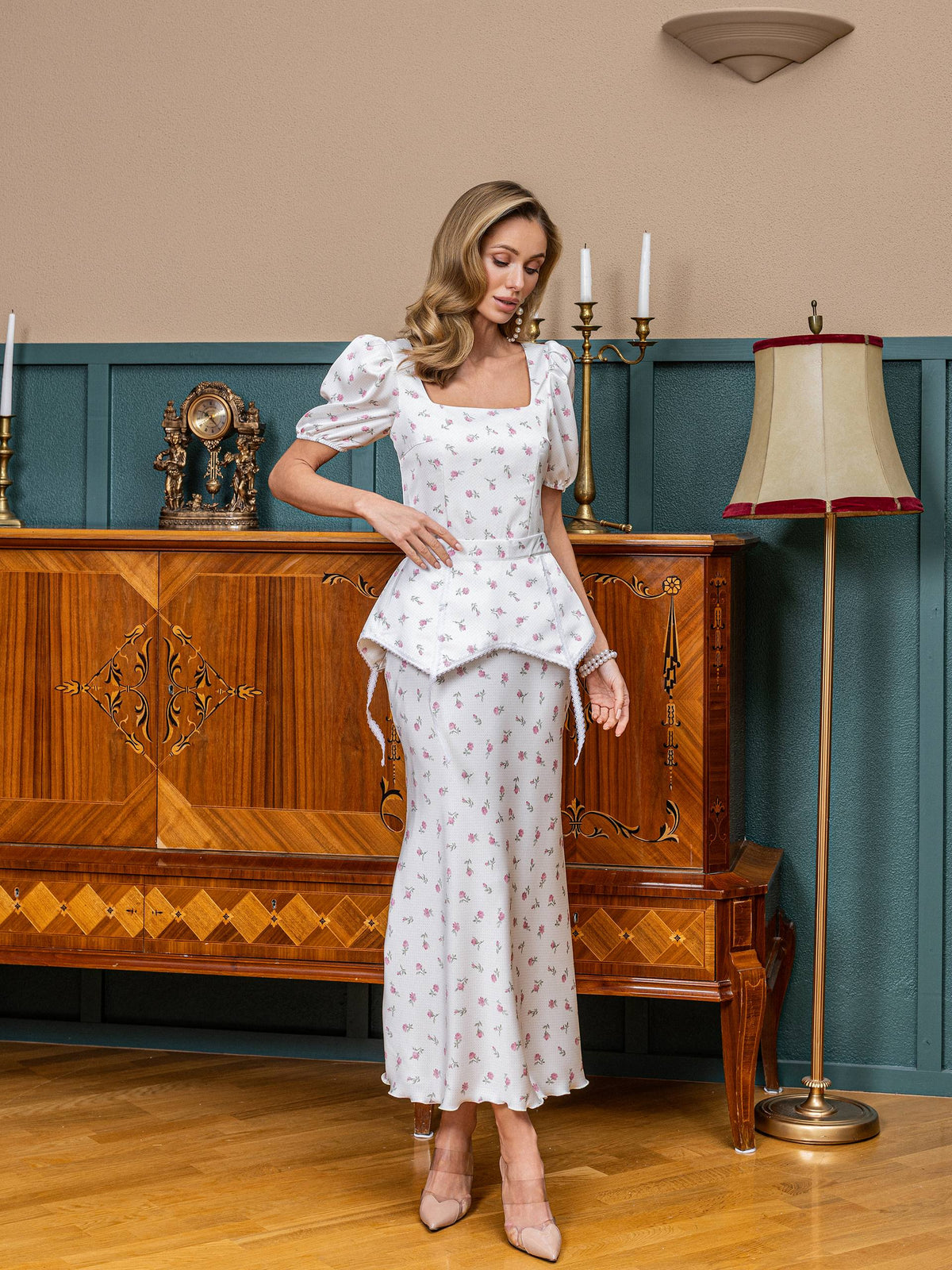 Woman in a white floral dress standing in a room with wooden furniture and decor.