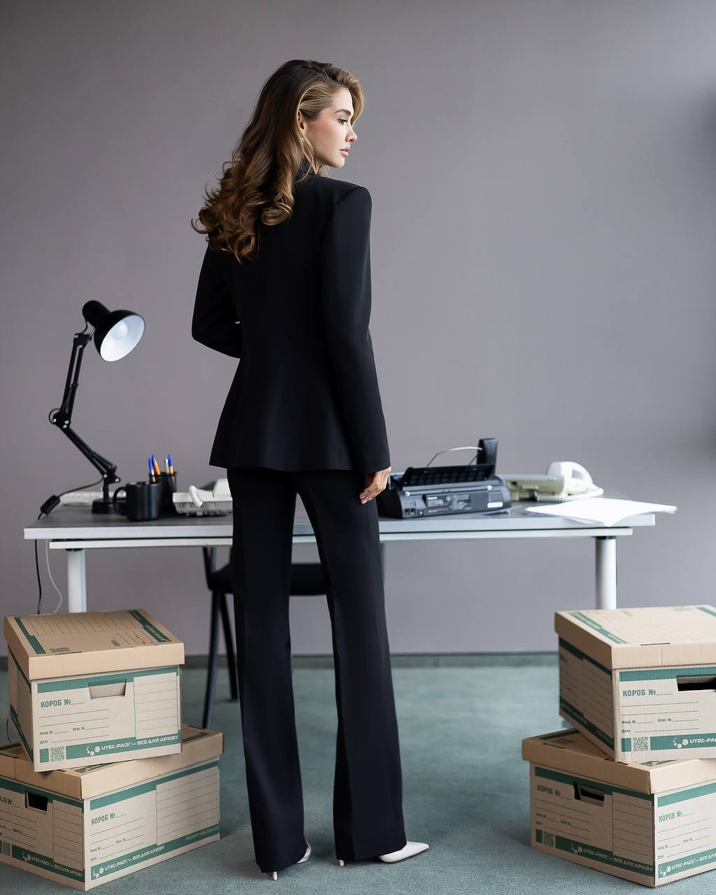 Woman in a black suit standing in an office with boxes and a desk.