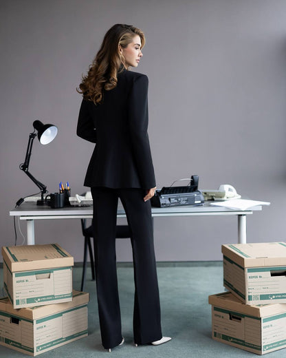 Woman in a black suit standing in an office with boxes and a desk.