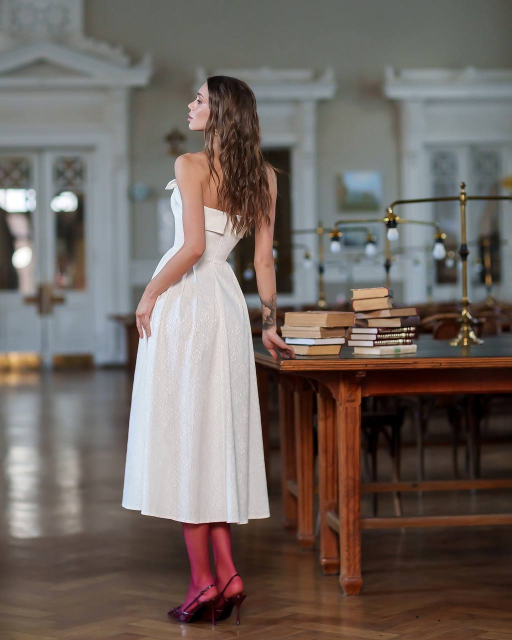 Woman in a white dress standing next to a table with books in a room with high ceilings and large windows.