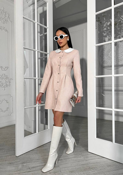 Woman in a light pink dress with white collar and white knee-high boots standing in a room with large glass doors.