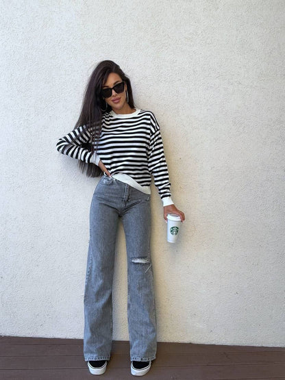 Woman wearing a black and white striped sweater and gray jeans, holding a Starbucks cup against a plain background.