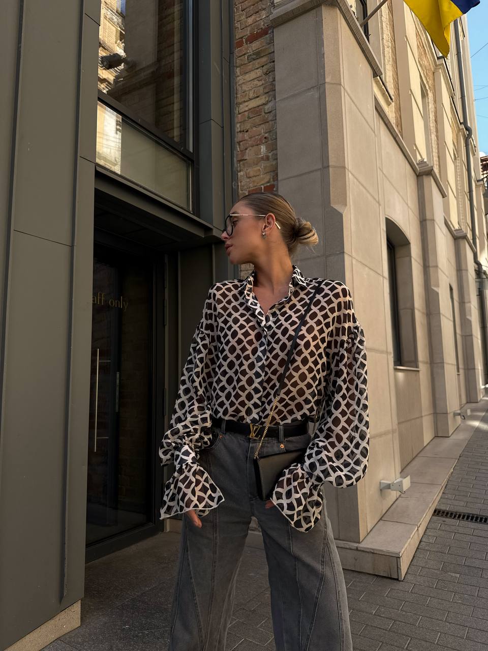 Woman in a patterned blouse and gray pants standing on a city street.