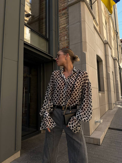 Woman in a patterned blouse and gray pants standing on a city street.