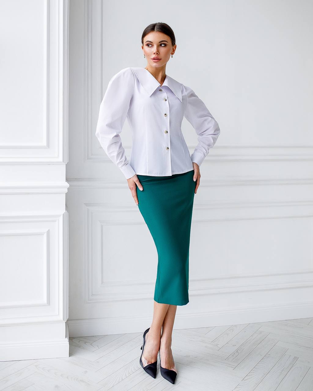 Woman wearing a white blouse and teal skirt against a white wall.