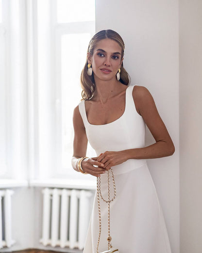 Woman in a white dress standing in a bright room with large windows.