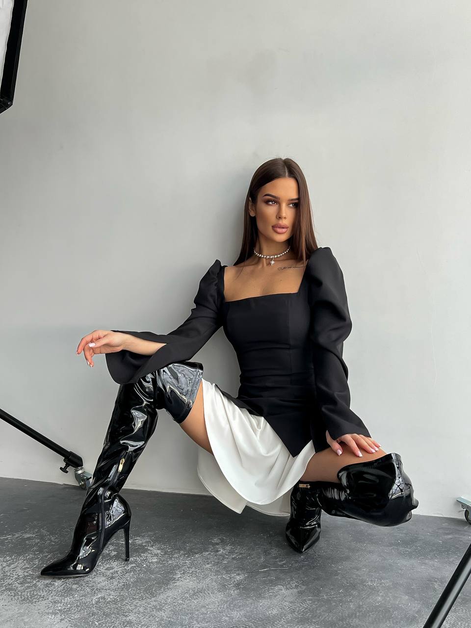 Woman in a black outfit with white skirt and black high-heeled boots posing against a gray background