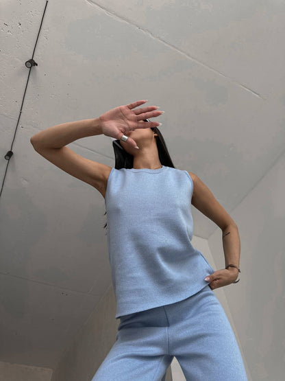 Person wearing a light blue sleeveless top and pants against a plain wall.