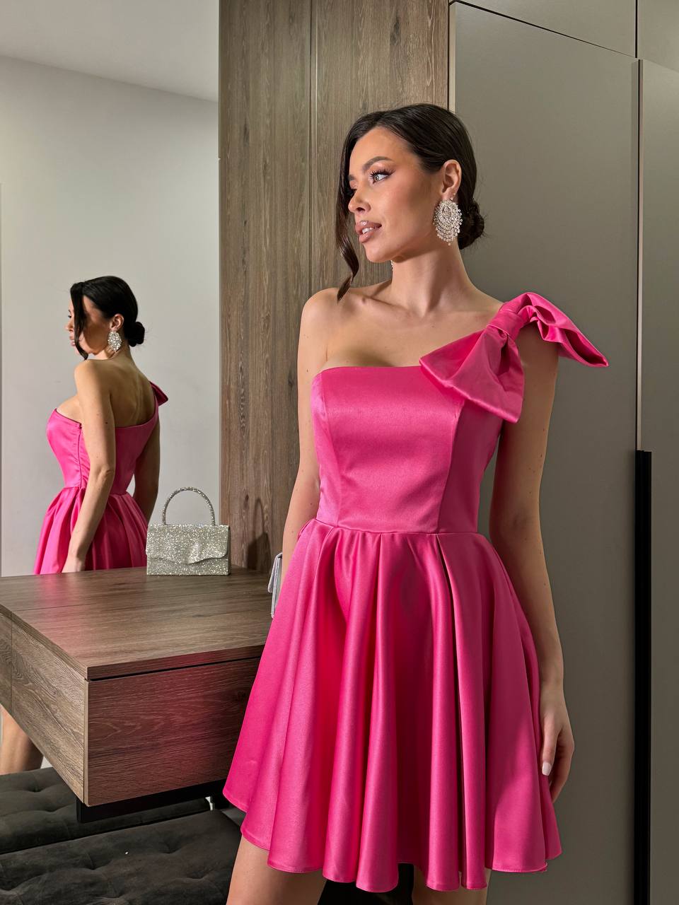 Woman wearing a pink one-shoulder dress with a bow detail, standing in front of a mirror.