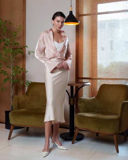 Woman in a pink jacket and white dress standing in a stylish room with green chairs and a lamp.