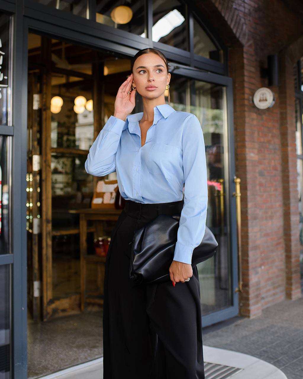 Woman wearing a light blue shirt and black pants standing in front of a store entrance.