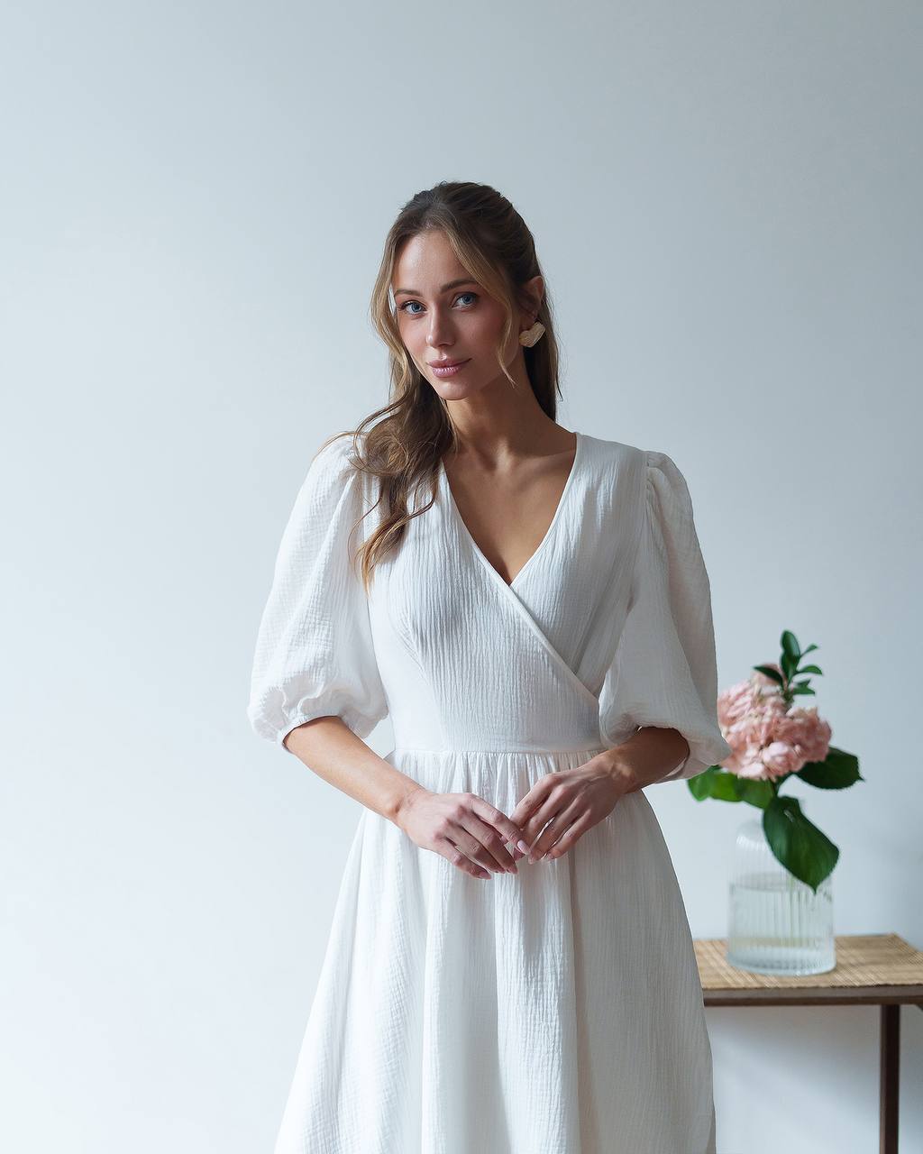 Woman wearing a white dress standing in a minimalistic room with a vase of flowers on a table.