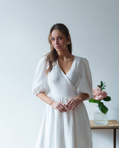 Woman wearing a white dress standing in a minimalistic room with a vase of flowers on a table.
