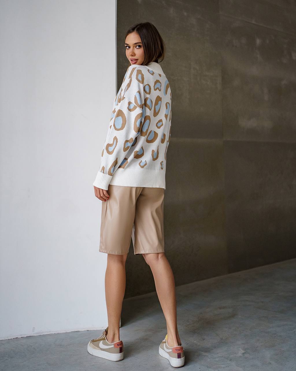 Woman wearing a patterned sweater and beige shorts against a plain wall.