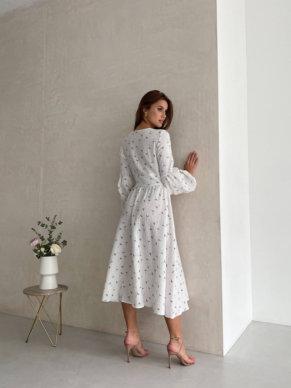 Woman in a white dress standing against a neutral wall with a small table and vase.